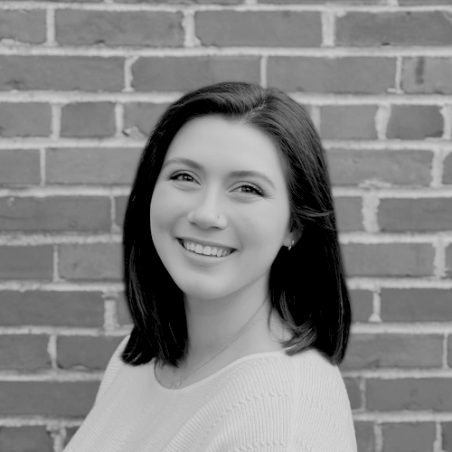 A woman with shoulder-length dark hair smiles at the camera while standing in front of a brick wall. The photo is in black and white.