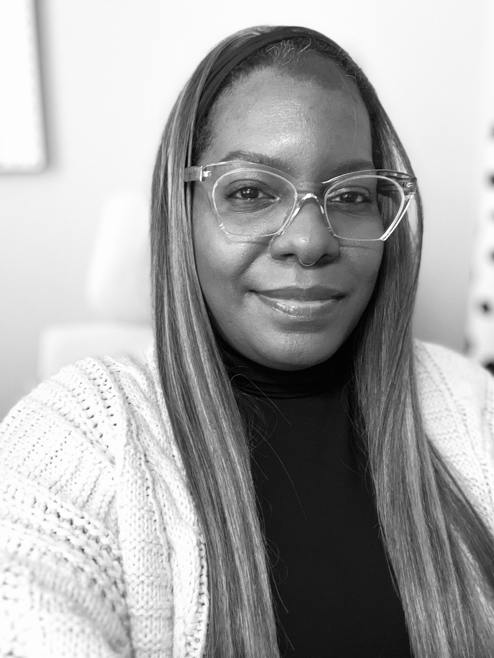 Black and white photo of a woman wearing glasses, a knit cardigan, and a turtleneck, looking at the camera and smiling slightly.