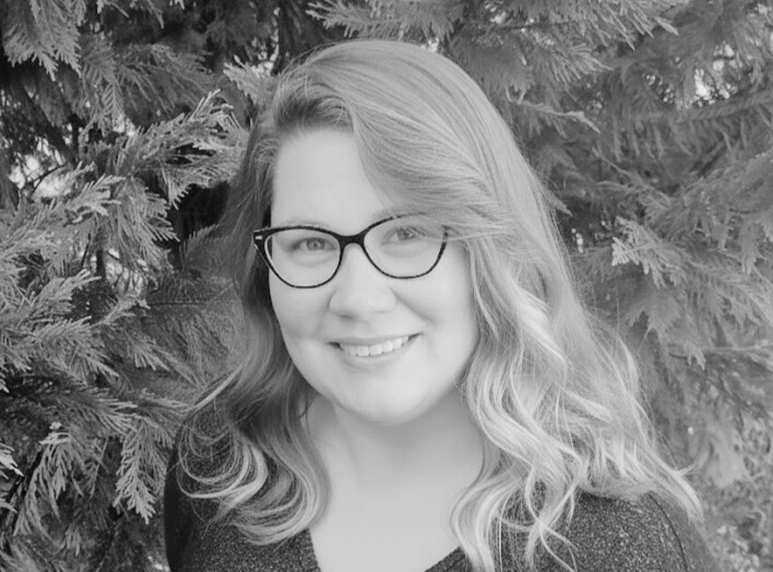 A woman with wavy hair and glasses smiles at the camera while standing in front of a tree with dense foliage. The image is in black and white.