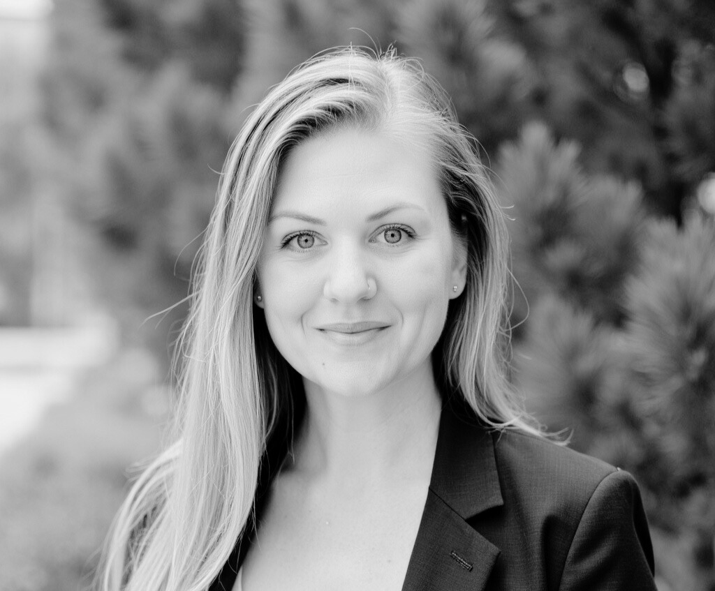 A woman with long hair wearing a blazer smiles at the camera outdoors, with trees blurred in the background. The image is in black and white.