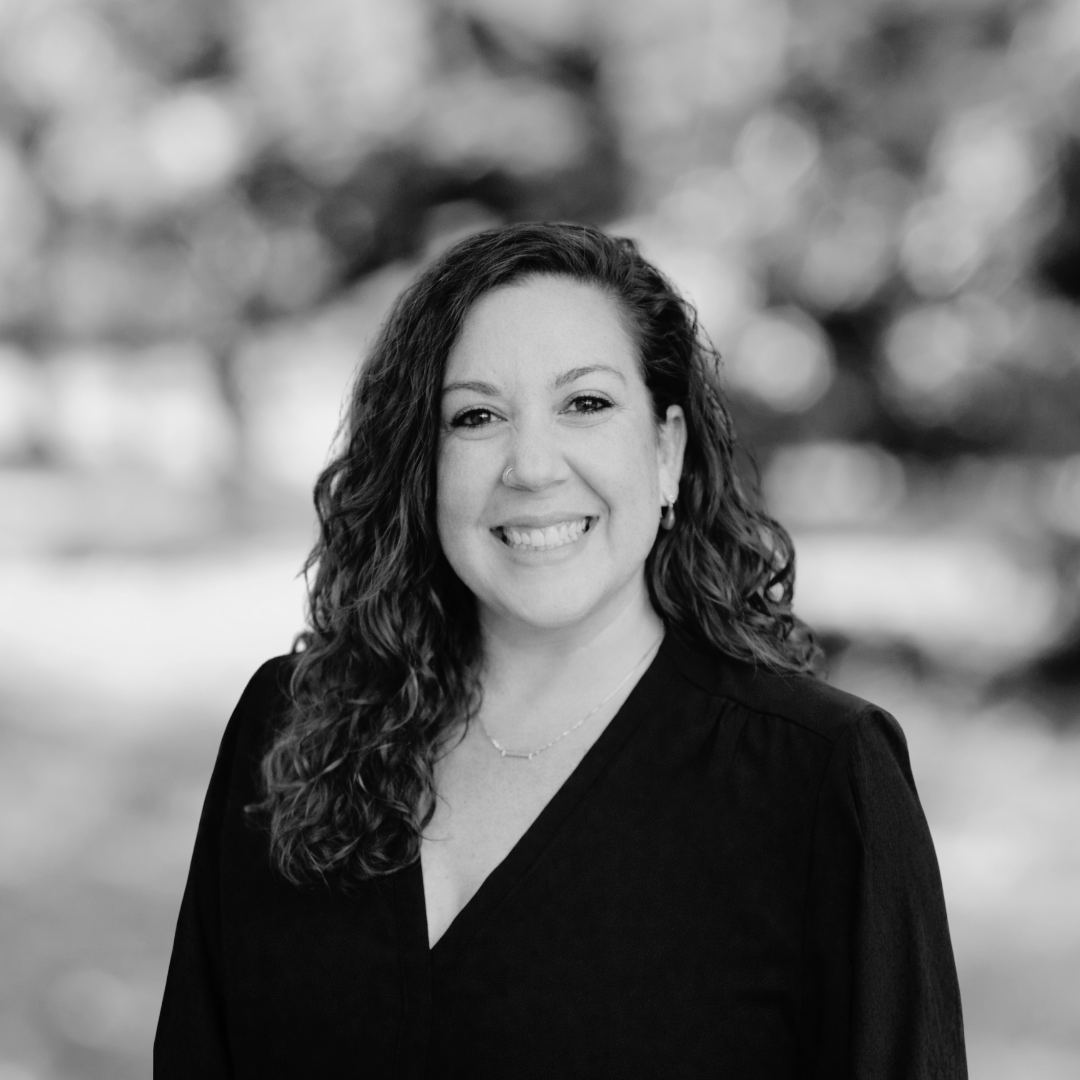 Woman with curly hair smiling, wearing a dark blouse, standing outdoors with a blurred background in black and white.