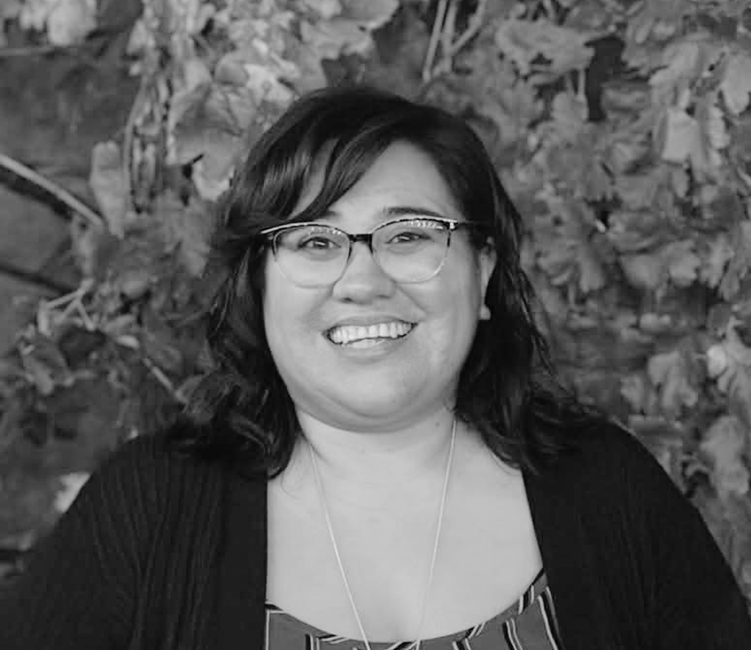 A woman with medium-length dark hair and glasses smiles at the camera, standing in front of leafy foliage. The image is in black and white.