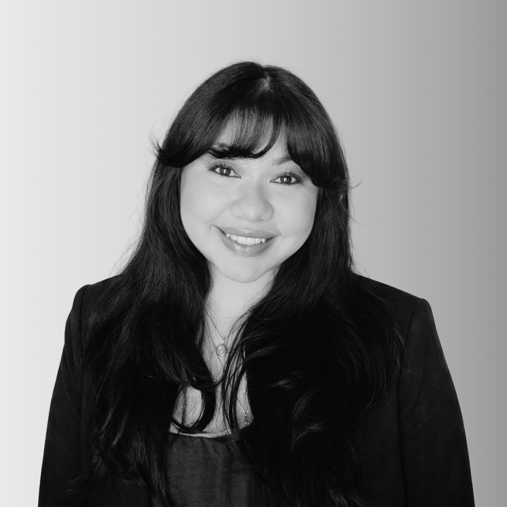 A woman with long dark hair and bangs, wearing a dark blazer and top, smiles at the camera against a gradient light gray background.