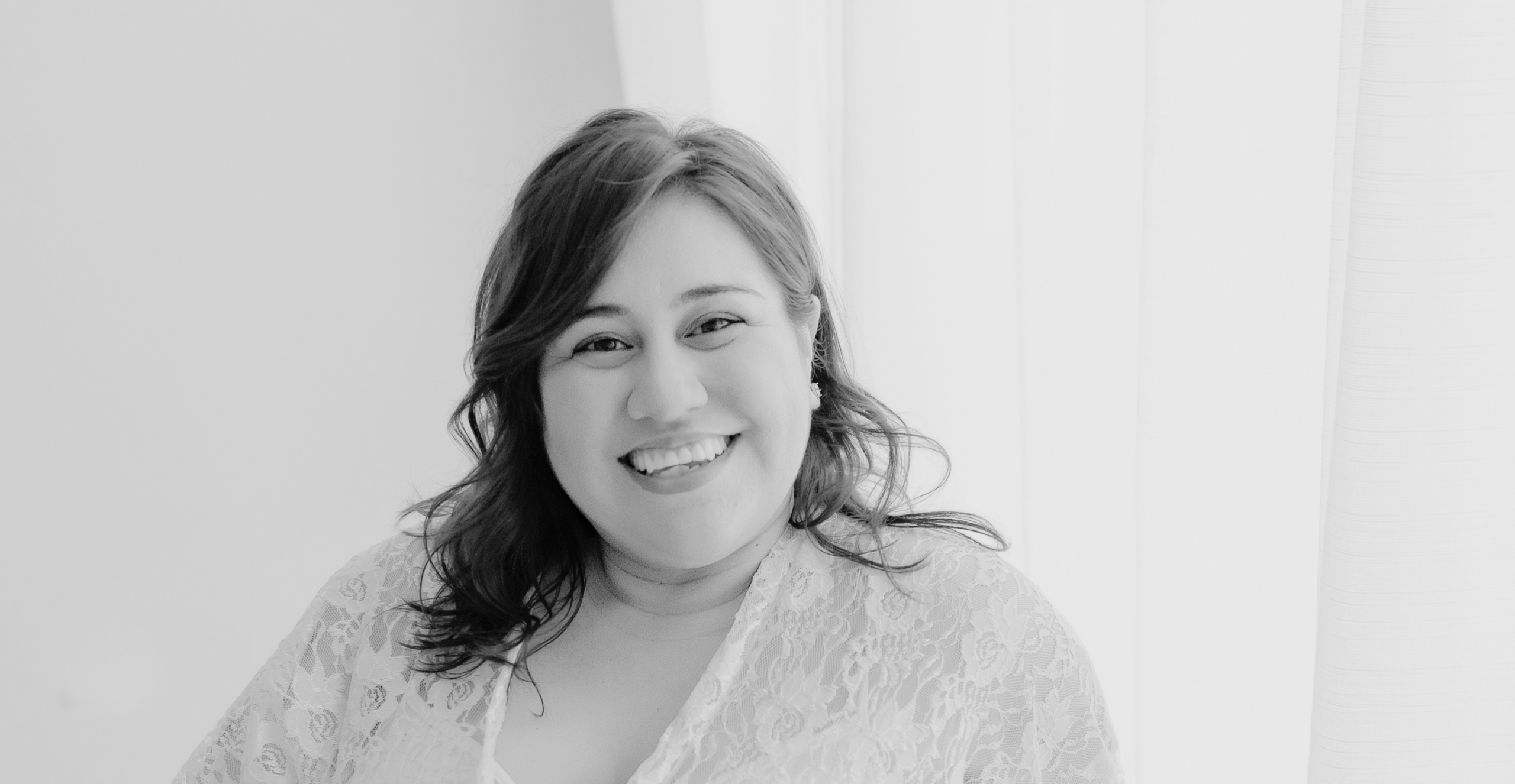 Woman with shoulder-length hair smiling, wearing a lace top, standing in front of a bright window in a softly lit room.