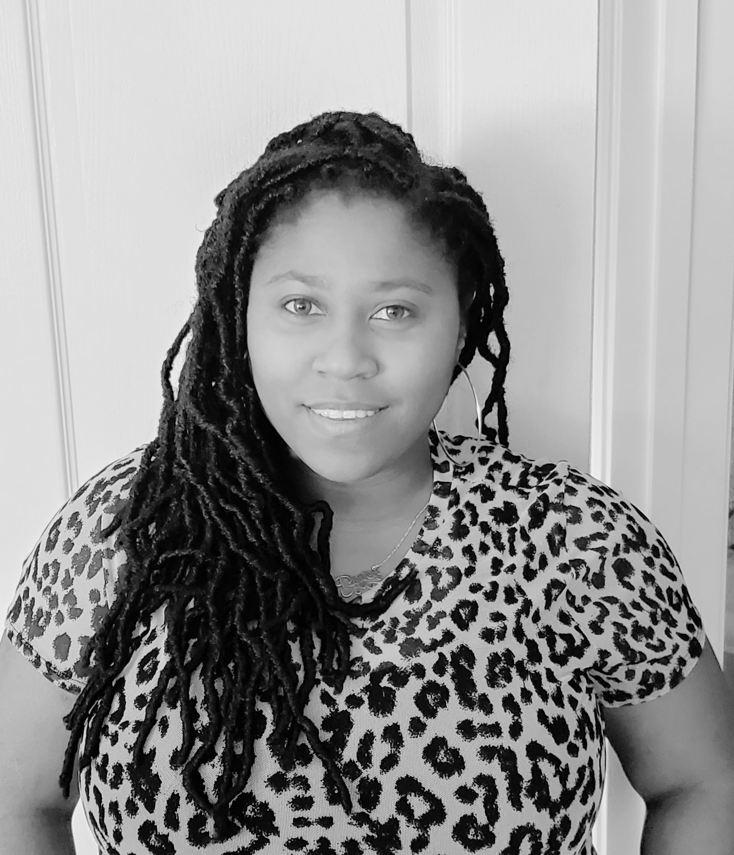 A woman with long, braided hair stands in front of a door, wearing a leopard print top and a necklace, looking at the camera and smiling slightly.