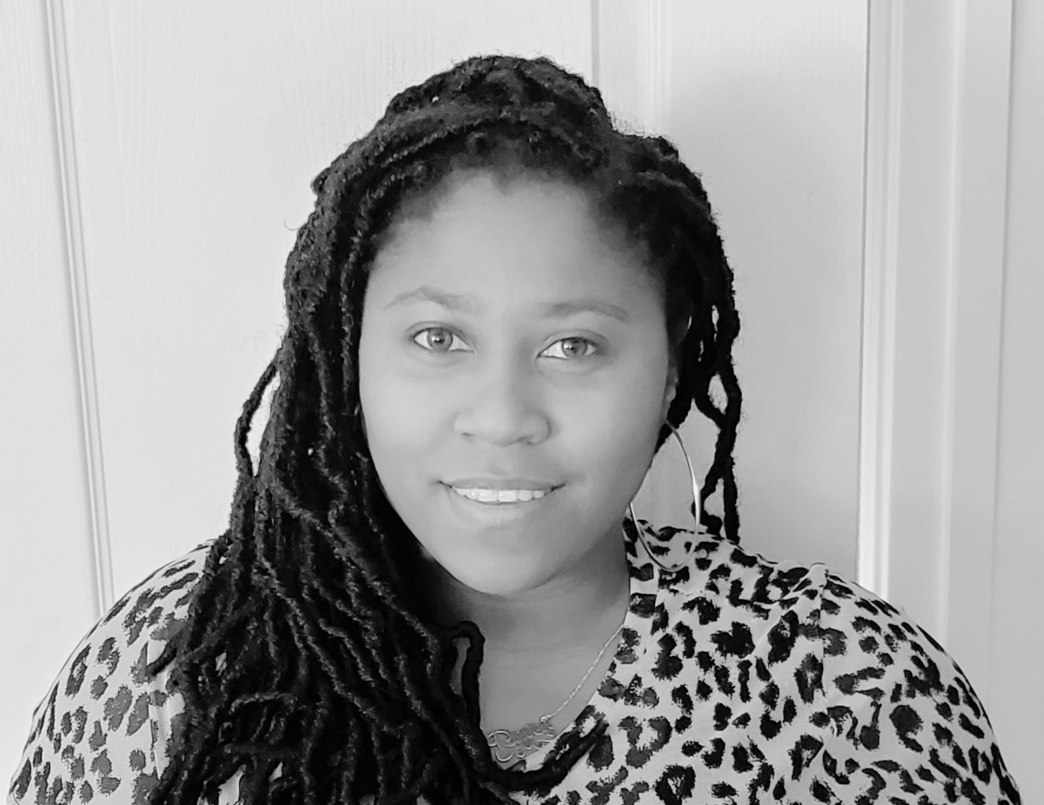 A woman with long, braided hair wearing a leopard print top stands in front of a light-colored wall, looking at the camera and smiling.