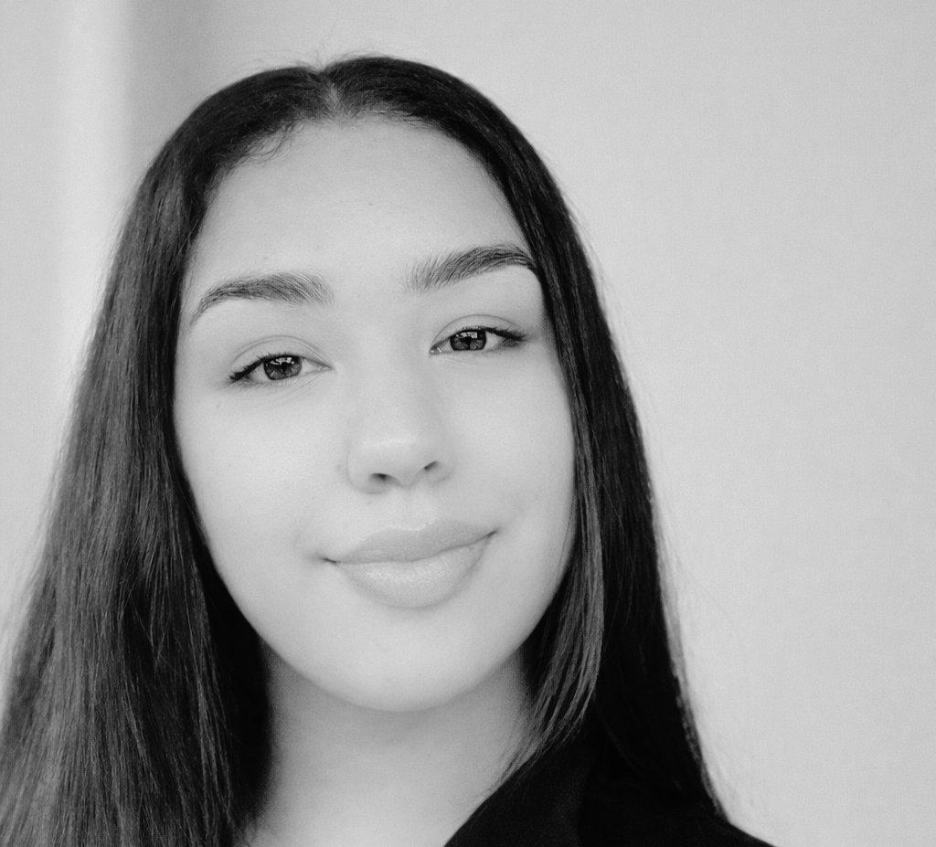 A young woman with long straight hair and a slight smile, photographed in black and white against a plain background.