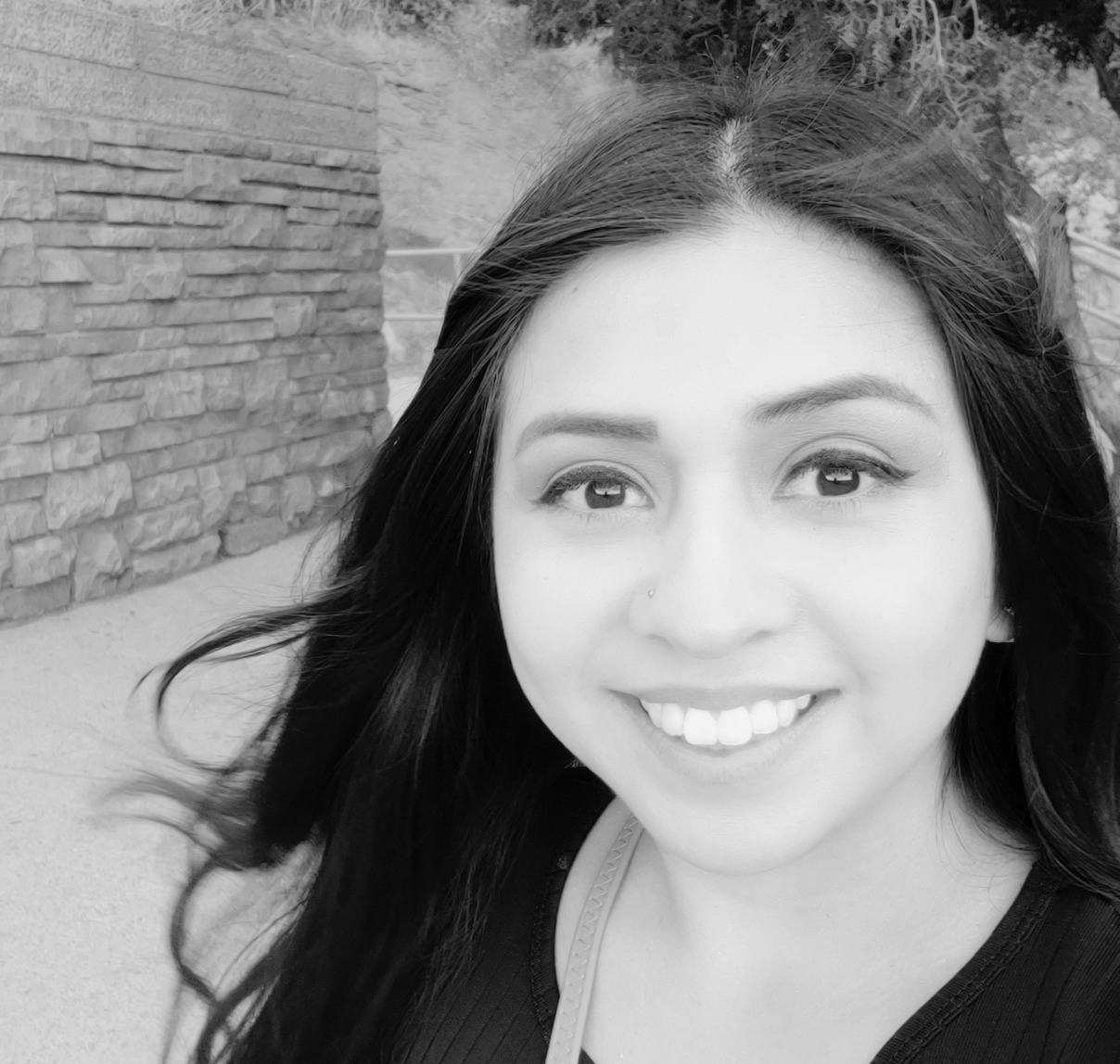 A woman with long dark hair smiles at the camera while standing outdoors near a stone wall.
