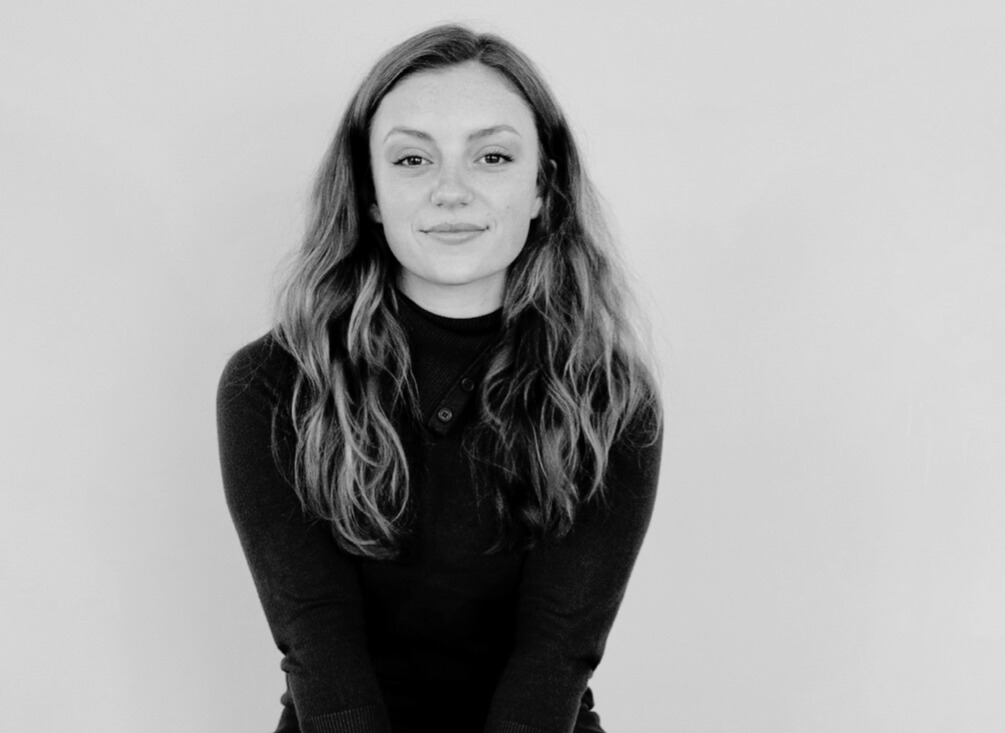 A black-and-white photo of a woman with long hair, wearing a dark long-sleeve top, seated against a plain background, looking at the camera.