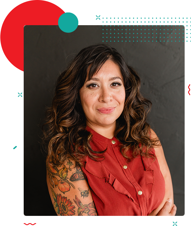 About a woman with wavy hair and an arm tattoo, standing in front of a dark gray wall, wearing a sleeveless red top.