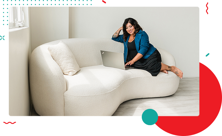 A woman sits on a white, curved sofa at home, using a laptop. She is dressed in a blue blazer and black dress, and the room has minimalistic decor.