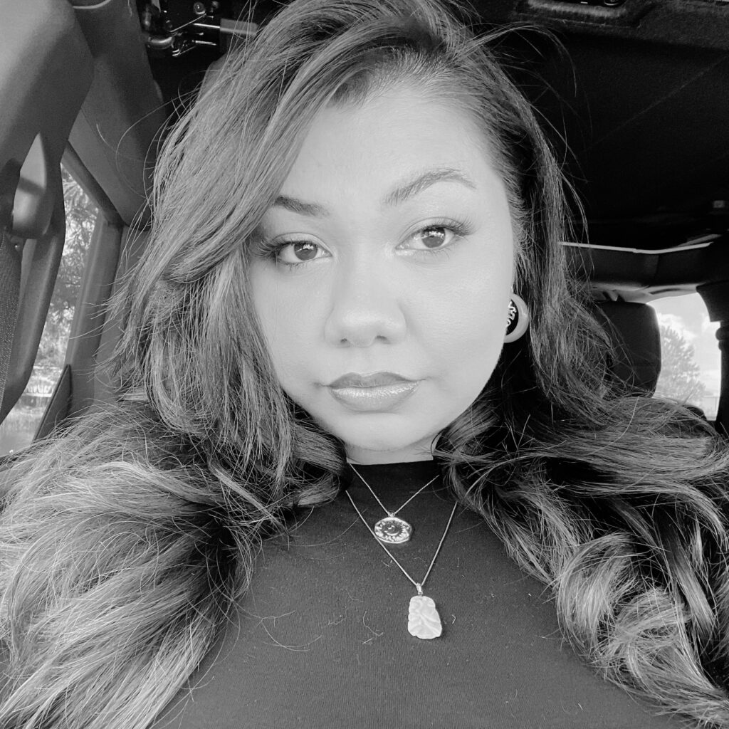 A woman with long hair sits in a car, looking at the camera. She is wearing a black top, hoop earrings, and two pendant necklaces. The image is in black and white.