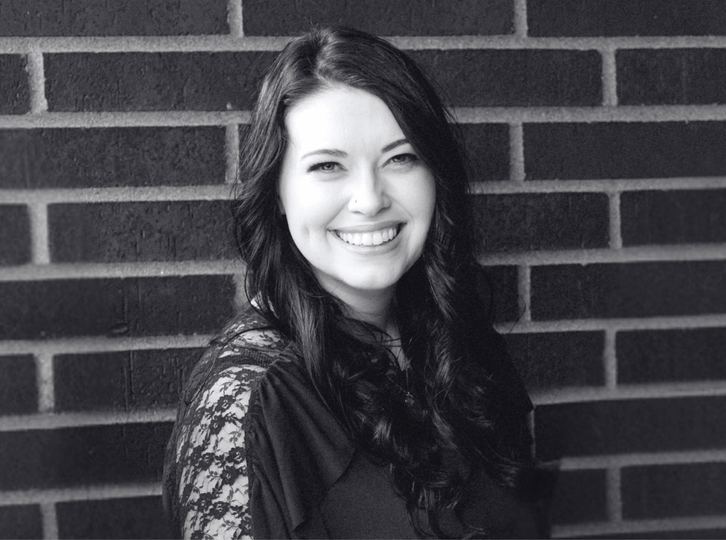 A woman with long dark hair smiles at the camera, standing in front of a brick wall. The photo is in black and white.
